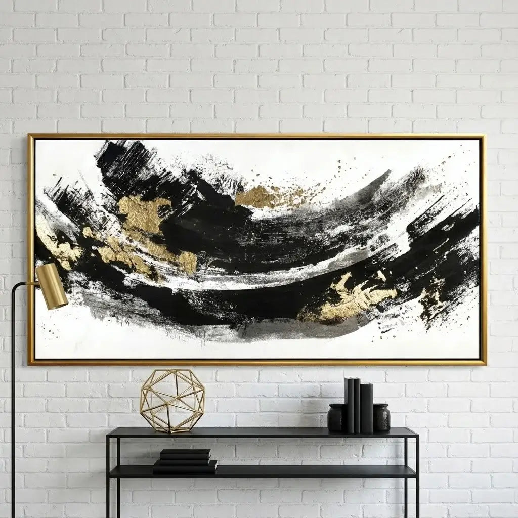 Abstract art piece with black and gold brush strokes in a gold frame on a white brick wall.