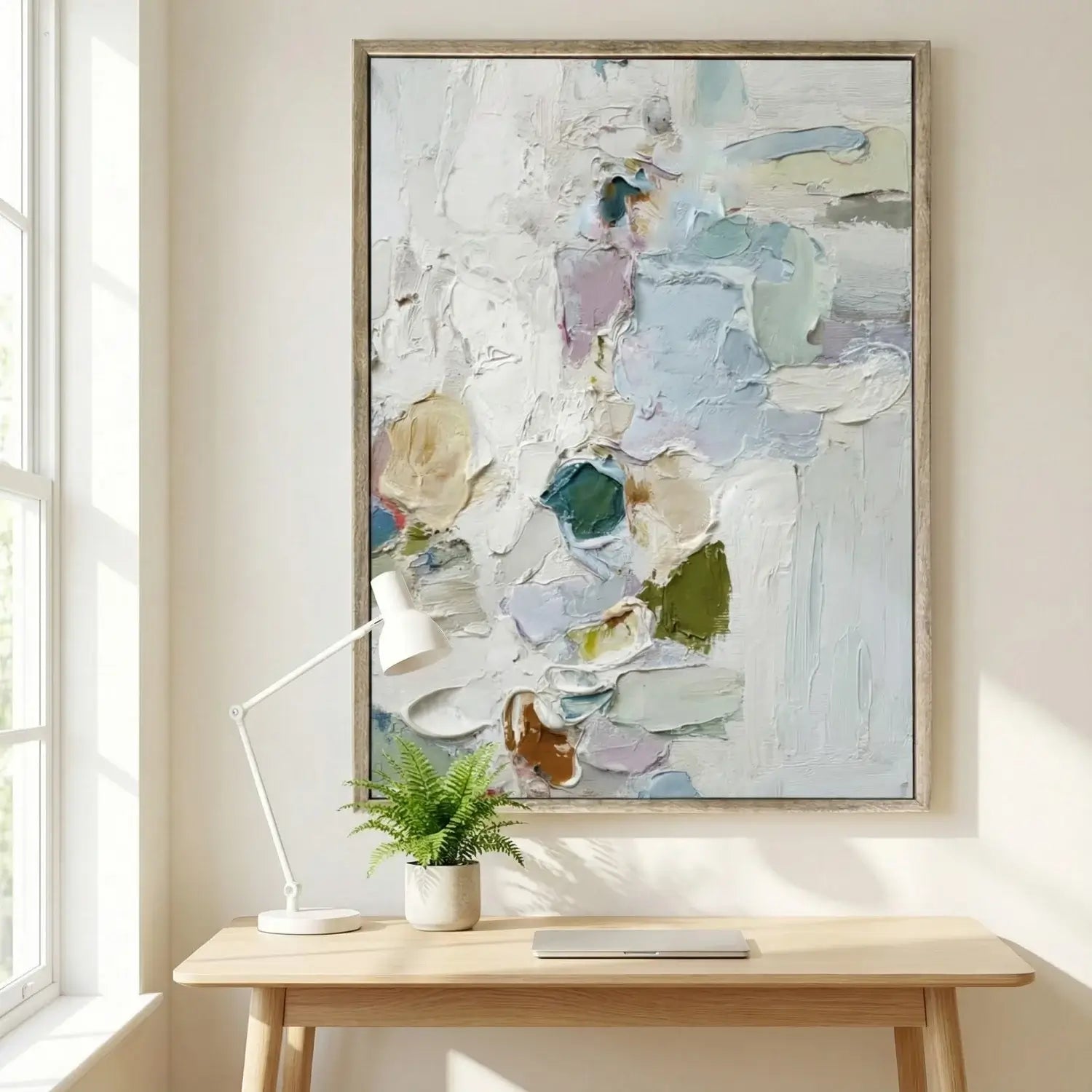 Abstract painting in a wooden frame on a wall above a desk with a lamp and plant.