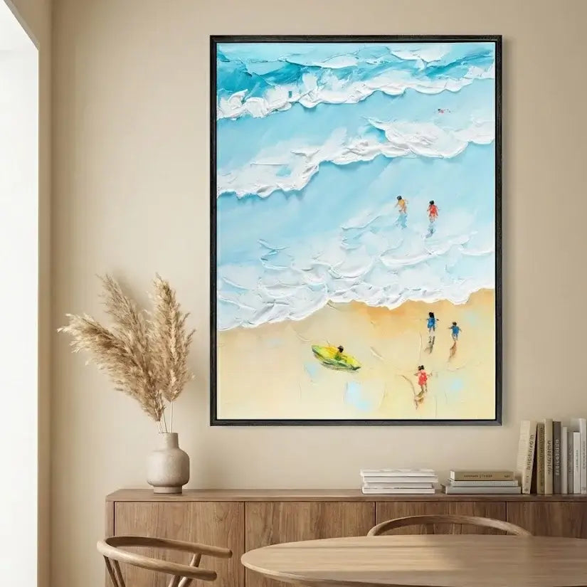 Framed artwork of people on a beach with a blue sky and white clouds, displayed in a room with a wooden cabinet and decor.
